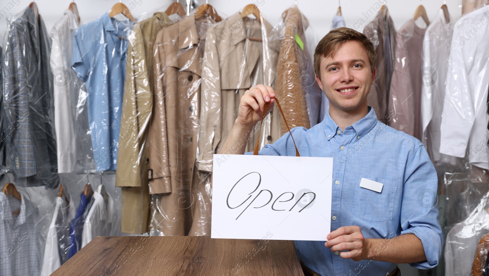 Photo of Dry-cleaning service. Happy worker holding Open sign at near rack with clothes indoors, space for text