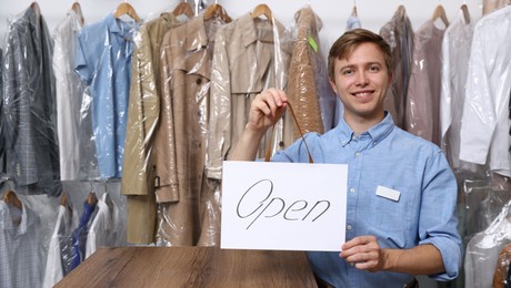 Photo of Dry-cleaning service. Happy worker holding Open sign at near rack with clothes indoors, space for text