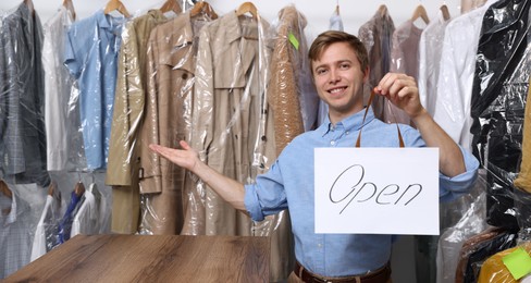 Photo of Dry-cleaning service. Happy worker holding Open sign at near rack with clothes indoors, space for text
