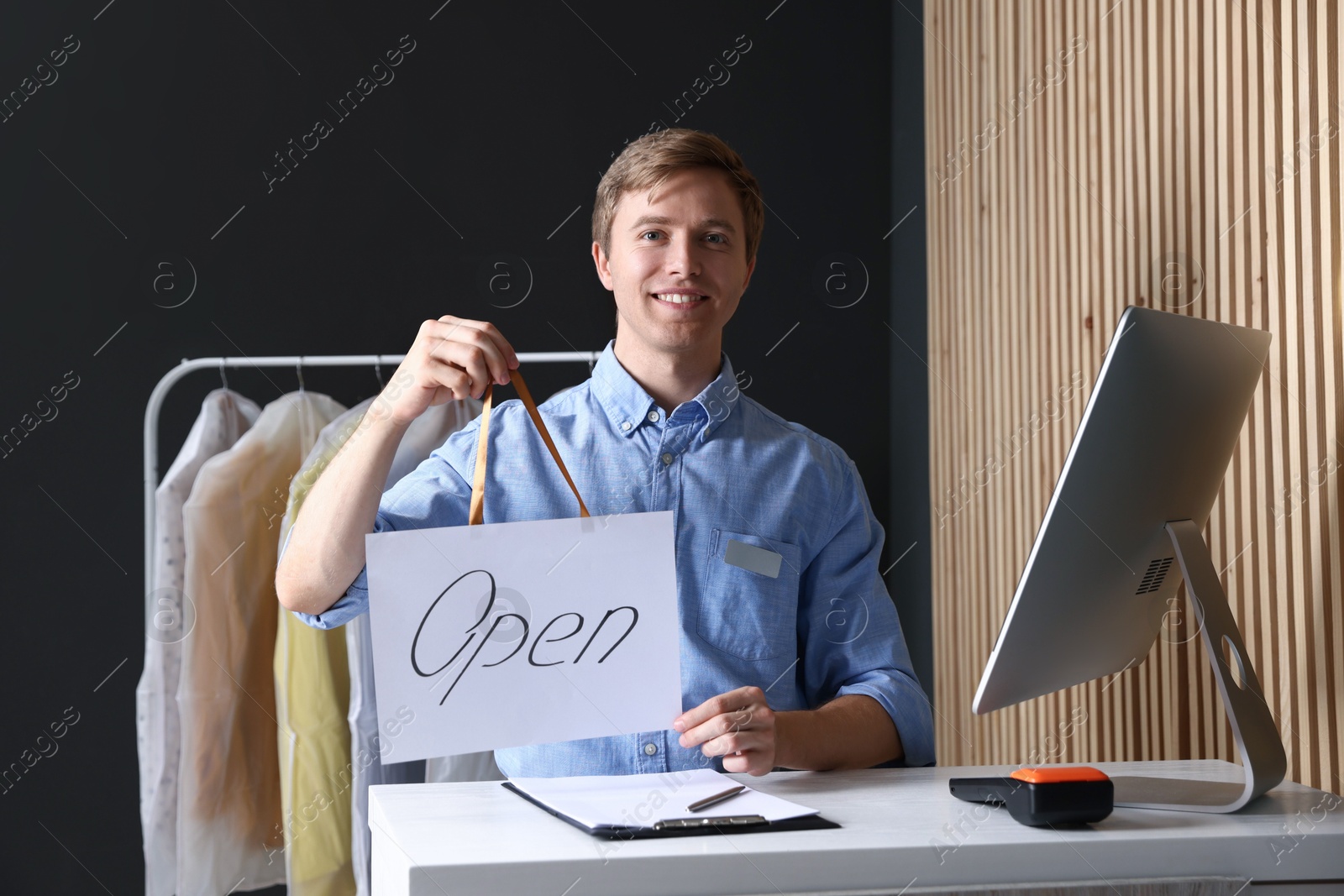 Photo of Dry-cleaning service. Happy worker holding Open sign at counter indoors