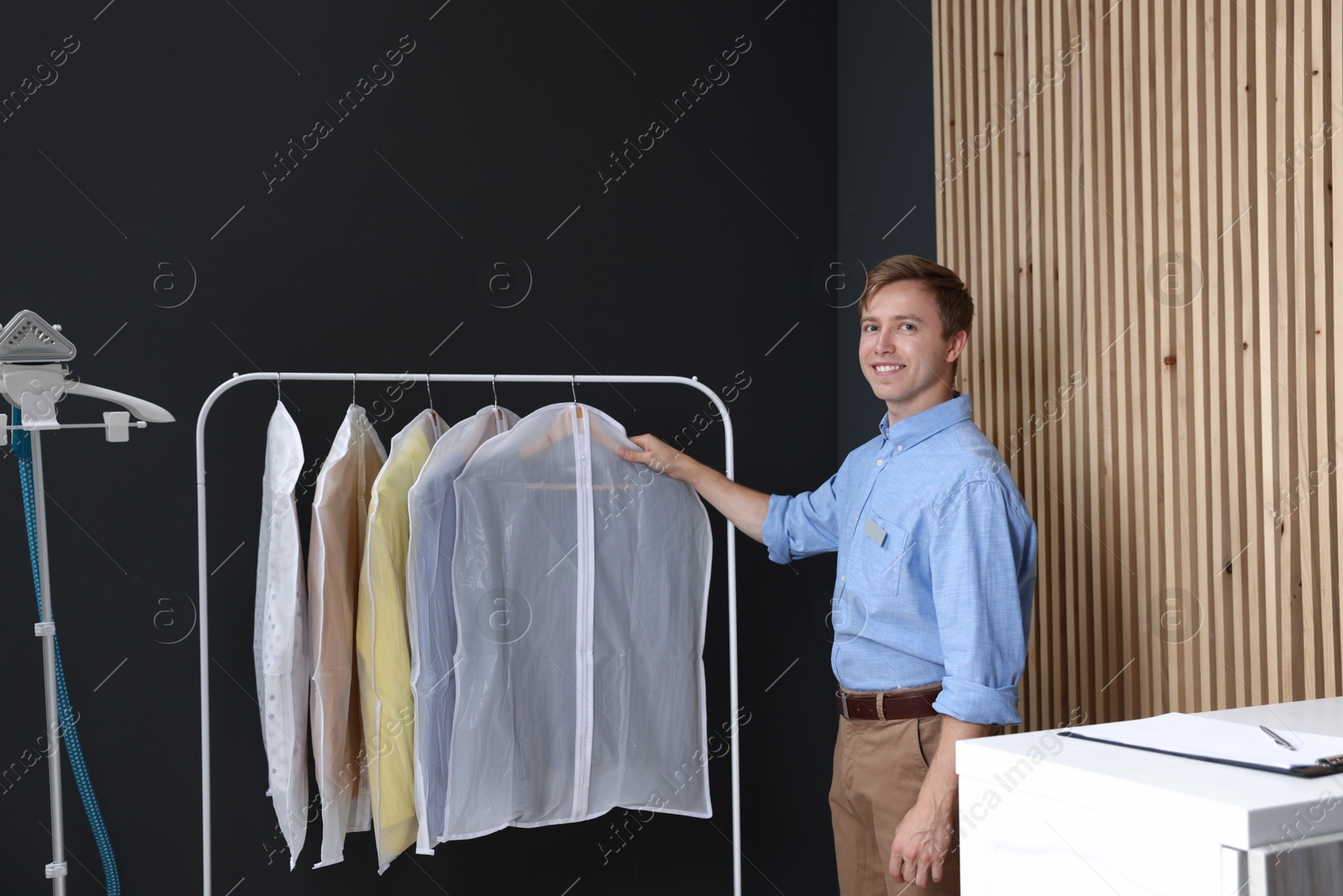 Photo of Dry-cleaning service. Happy worker near rack with clothes in garment covers indoors
