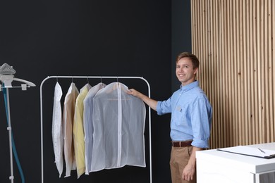 Photo of Dry-cleaning service. Happy worker near rack with clothes in garment covers indoors