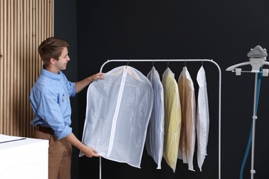 Photo of Dry-cleaning service. Happy worker choosing clothes from rack indoors