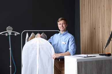 Photo of Dry-cleaning service. Happy worker holding shirt in garment cover indoors