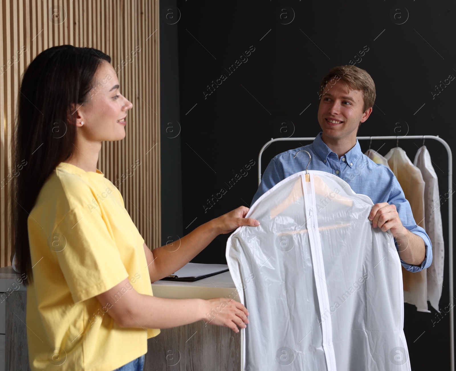Photo of Dry-cleaning service. Happy worker giving shirt in garment cover to client indoors