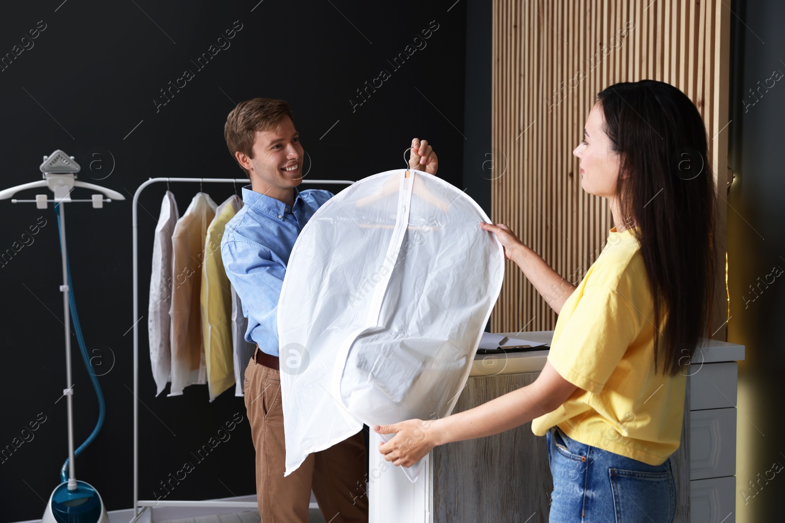 Photo of Dry-cleaning service. Happy worker giving shirt in garment cover to client indoors