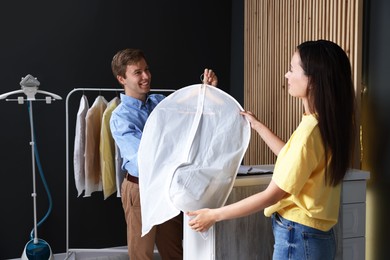 Photo of Dry-cleaning service. Happy worker giving shirt in garment cover to client indoors