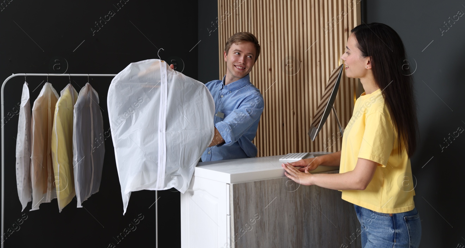 Photo of Dry-cleaning service. Happy worker giving shirt in garment cover to client indoors