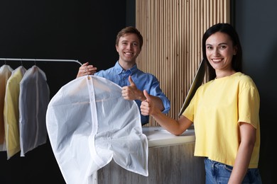 Photo of Dry-cleaning service. Happy worker with garment cover and client showing thumbs up indoors