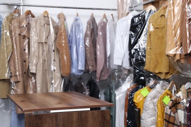 Photo of Dry-cleaning service. Many different clothes in plastic bags on rack and wooden counter indoors