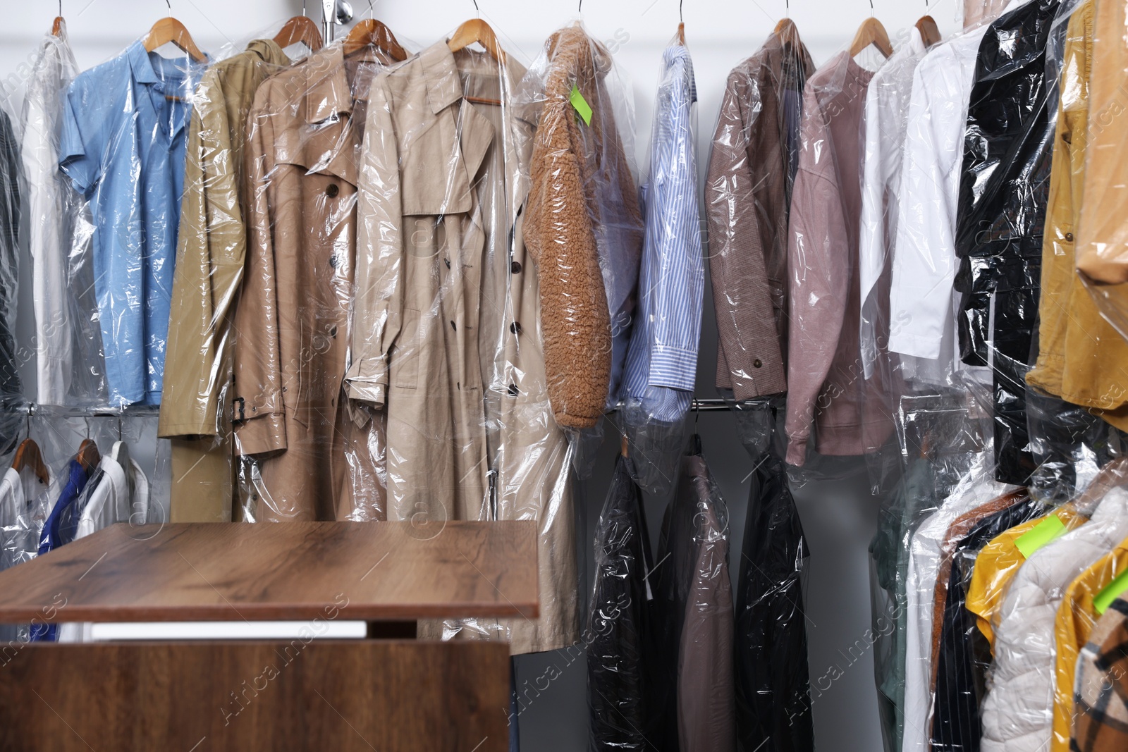 Photo of Dry-cleaning service. Many different clothes in plastic bags on rack and wooden counter indoors