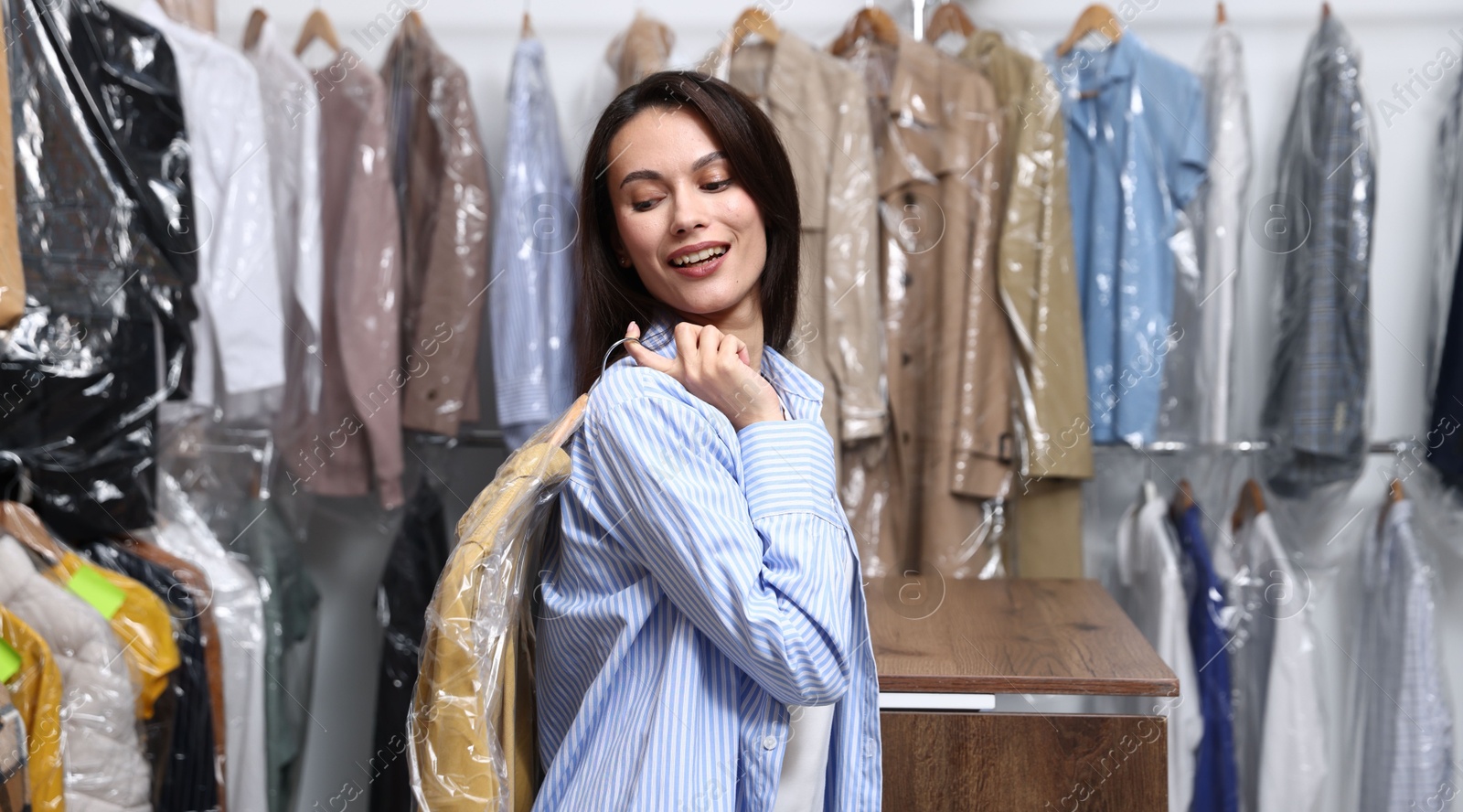 Photo of Dry-cleaning service. Happy woman holding hanger with shirt in plastic bag indoors