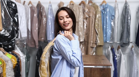 Photo of Dry-cleaning service. Happy woman holding hanger with shirt in plastic bag indoors