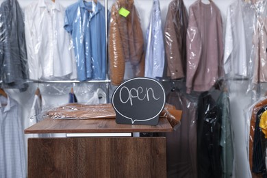 Photo of Dry-cleaning service. Open sign and jacket with plastic bag on wooden counter indoors