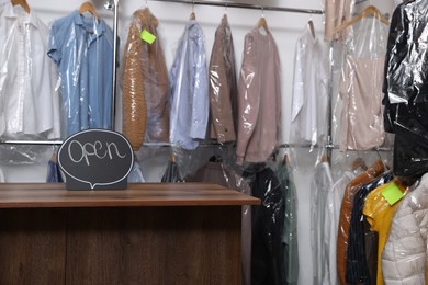Photo of Dry-cleaning service. Open sign on wooden counter near rack with clothes indoors