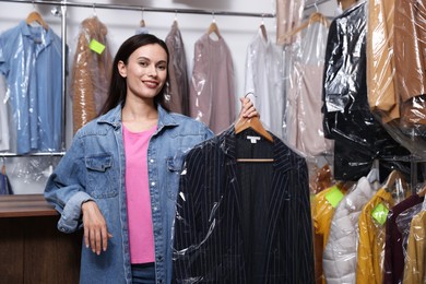 Photo of Dry-cleaning service. Happy woman holding hanger with jacket in plastic bag indoors