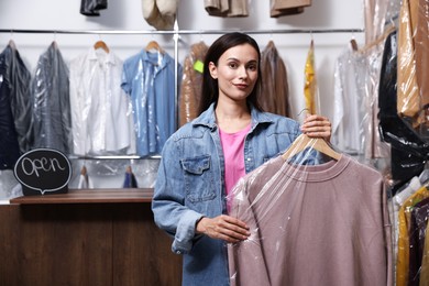 Photo of Dry-cleaning service. Happy woman holding hanger with sweatshirt in plastic bag indoors