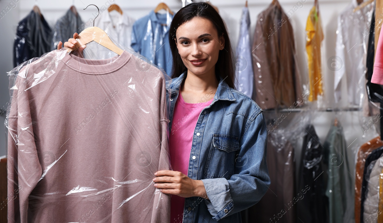 Dry-cleaning service. Happy woman holding hanger with sweatshirt in plastic bag indoors Photo of Dry-cleaning service. Happy woman holding hanger with sweatshirt in plastic bag indoors