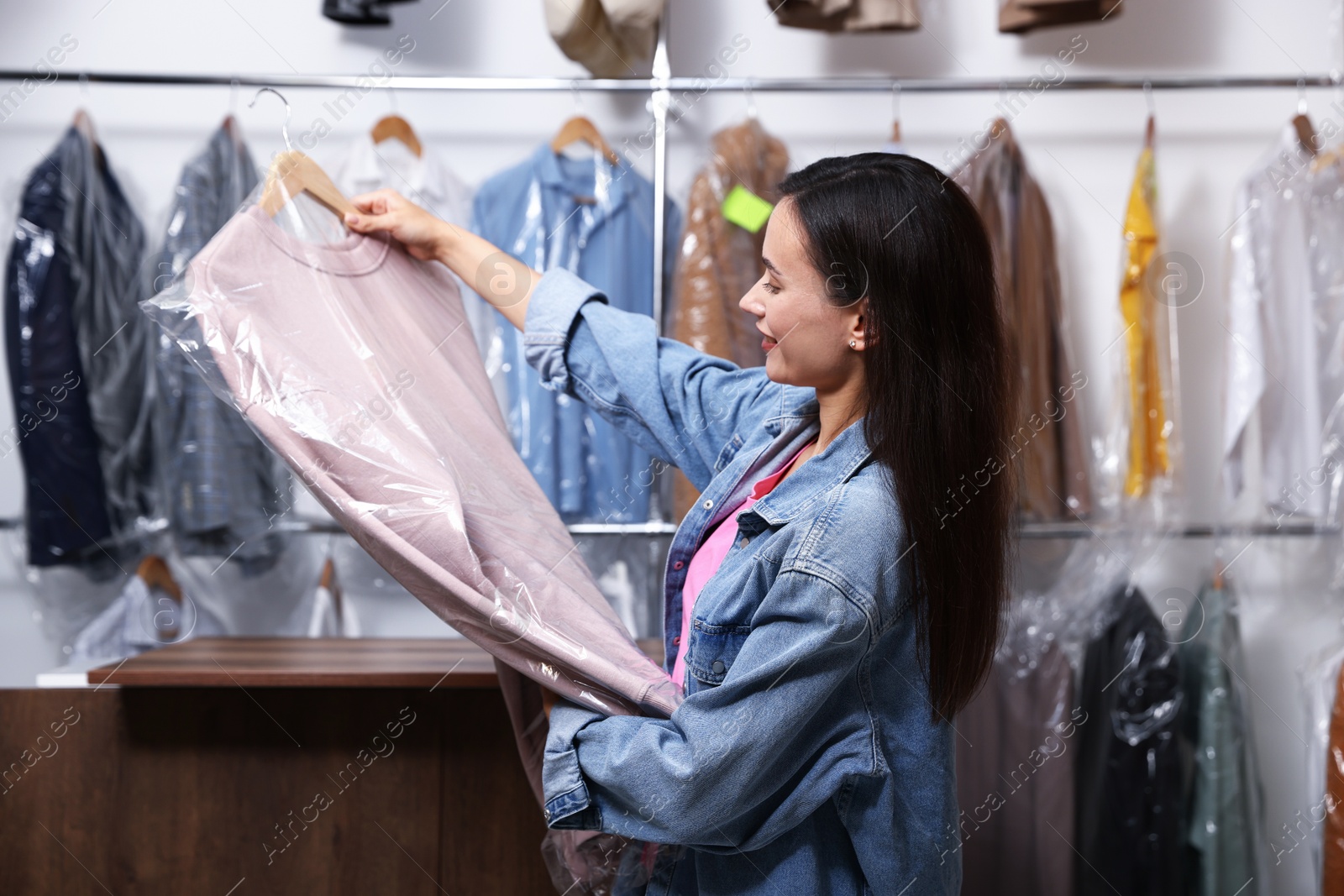 Photo of Dry-cleaning service. Happy woman holding hanger with sweatshirt in plastic bag indoors