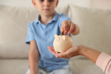 Photo of Mother and her son putting pocket money into piggy bank at home, closeup
