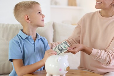 Photo of Mother and her son putting pocket money into piggy bank at wooden table indoors, closeup