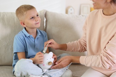 Photo of Mother and her son putting pocket money into piggy bank at home, closeup