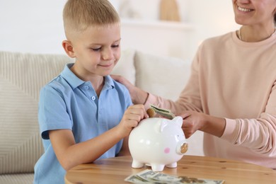 Photo of Mother and her son putting pocket money into piggy bank at wooden table indoors, closeup