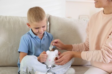 Photo of Mother and her son putting pocket money into piggy bank at home, closeup