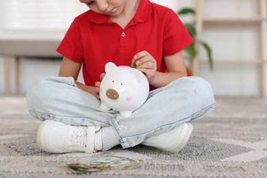 Photo of Little boy putting pocket money into piggy bank on floor at home, closeup
