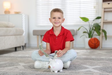 Photo of Little boy putting pocket money into piggy bank on floor at home
