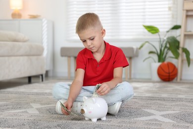 Photo of Little boy putting pocket money into piggy bank on floor at home