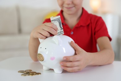 Photo of Little boy putting pocket money into piggy bank at white table indoors, closeup