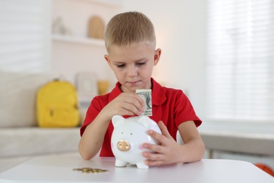 Photo of Little boy putting pocket money into piggy bank at white table indoors