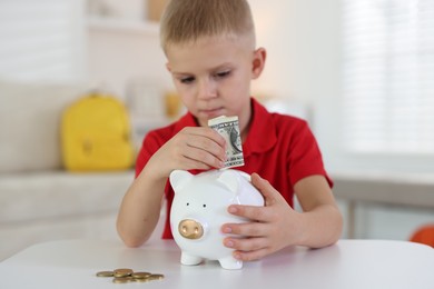 Photo of Little boy putting pocket money into piggy bank at white table indoors