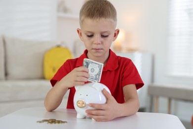 Photo of Little boy putting pocket money into piggy bank at white table indoors