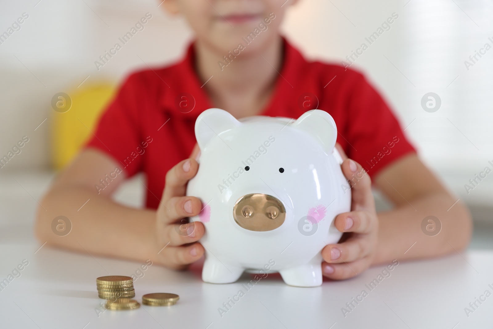 Photo of Little boy putting pocket money into piggy bank at white table indoors, closeup