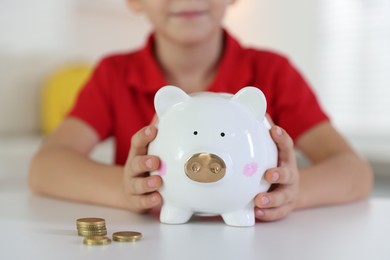 Photo of Little boy putting pocket money into piggy bank at white table indoors, closeup
