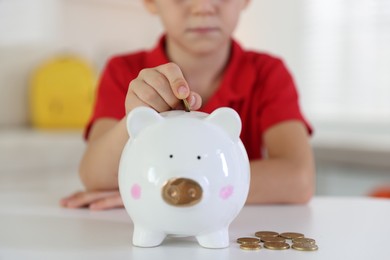 Photo of Little boy putting pocket money into piggy bank at white table indoors, closeup