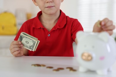 Photo of Little boy putting pocket money into piggy bank at white table indoors, closeup