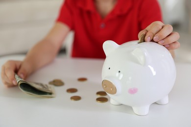 Photo of Little boy putting pocket money into piggy bank at white table indoors, closeup