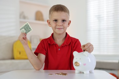 Photo of Little boy putting pocket money into piggy bank at white table indoors