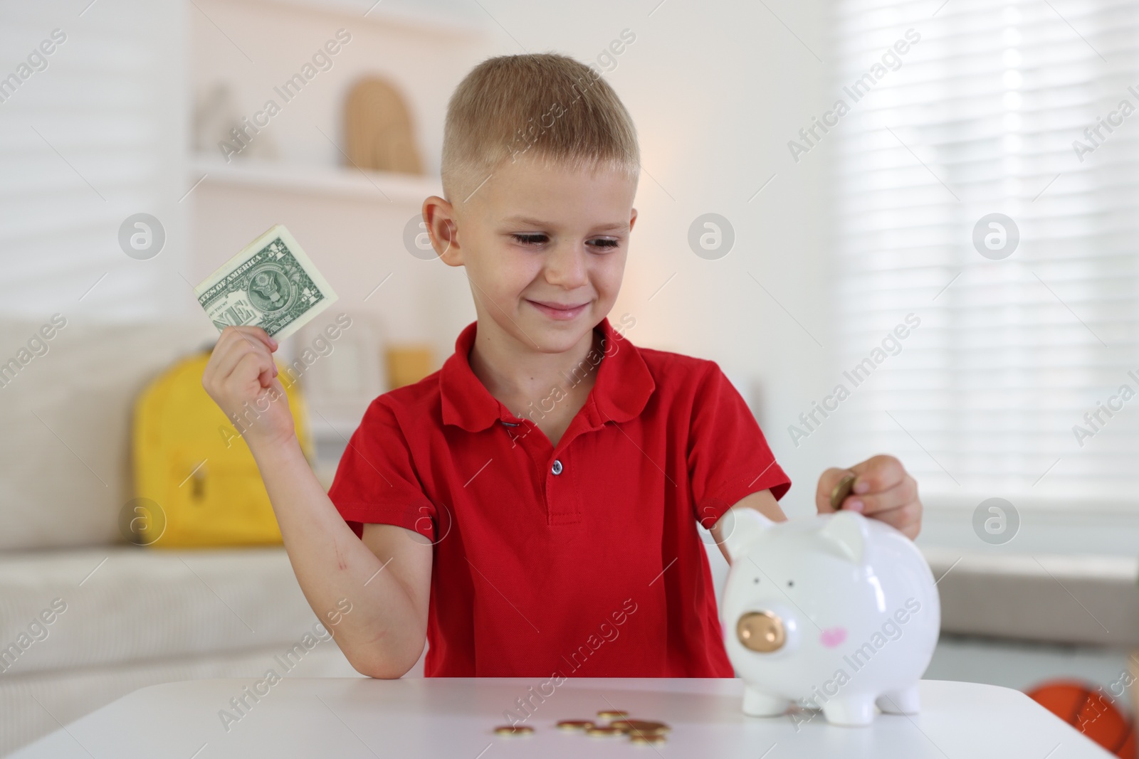 Photo of Little boy putting pocket money into piggy bank at white table indoors