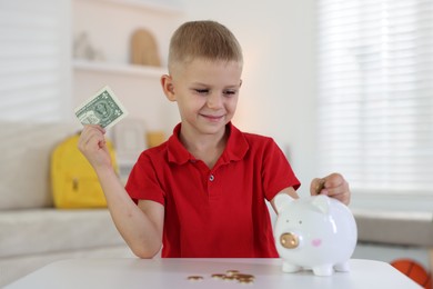 Photo of Little boy putting pocket money into piggy bank at white table indoors