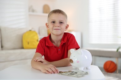 Photo of Little boy with pocket money and piggy bank at white table indoors
