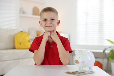 Photo of Little boy with pocket money and piggy bank at white table indoors