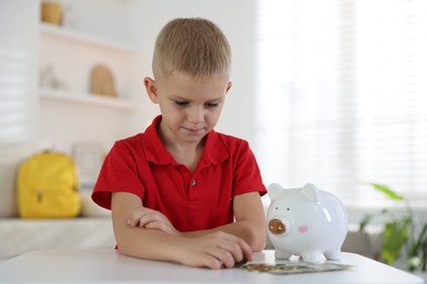 Photo of Little boy with pocket money and piggy bank at white table indoors
