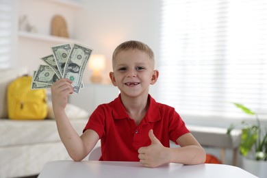 Photo of Little boy holding pocket money and showing thumbs up at white table indoors