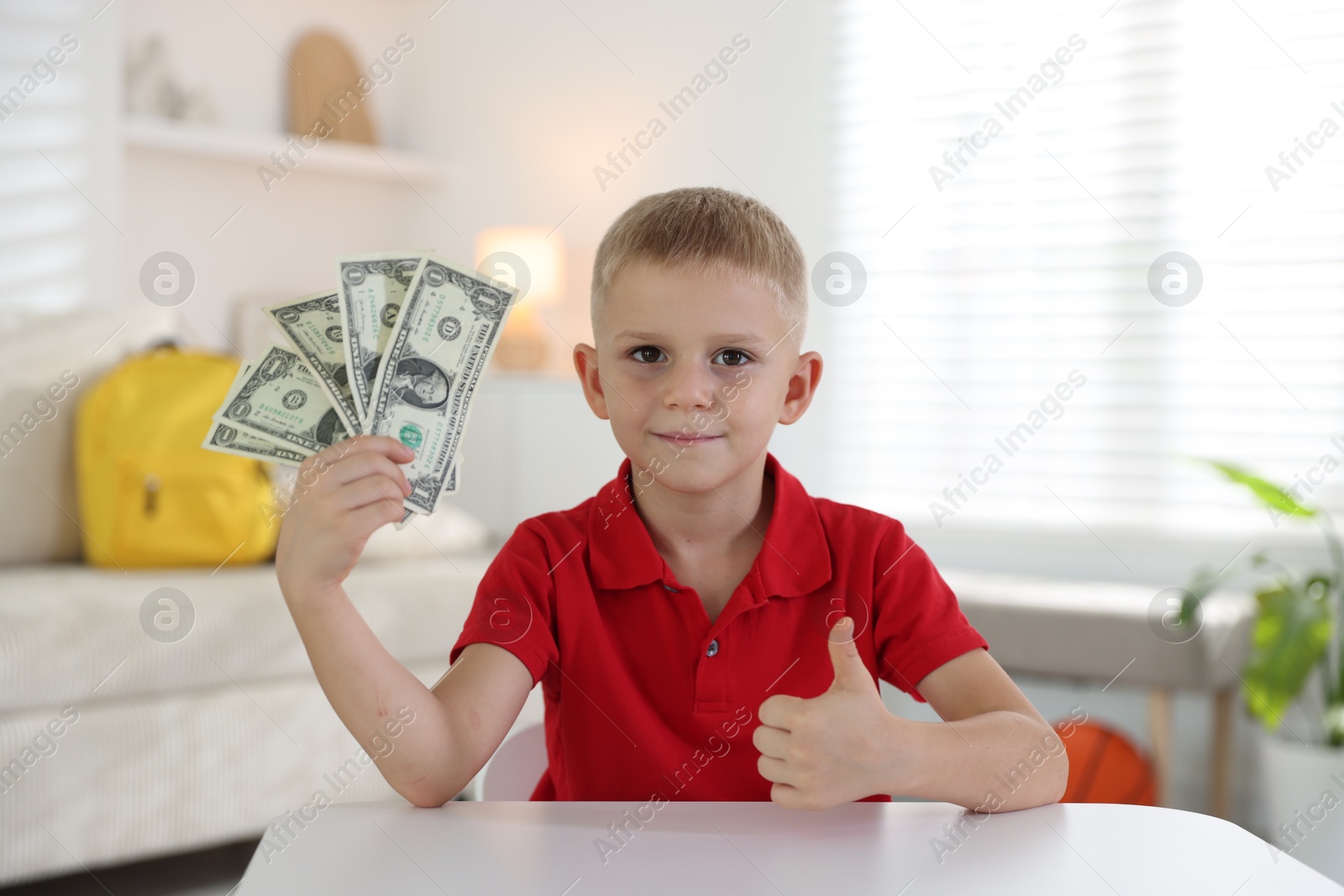 Photo of Little boy holding pocket money and showing thumbs up at white table indoors