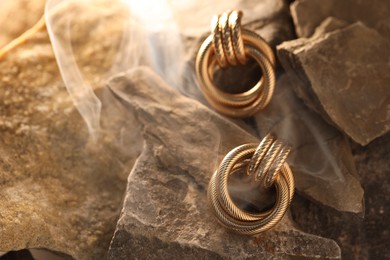Photo of Stylish gold earrings in smoke on rocks, closeup