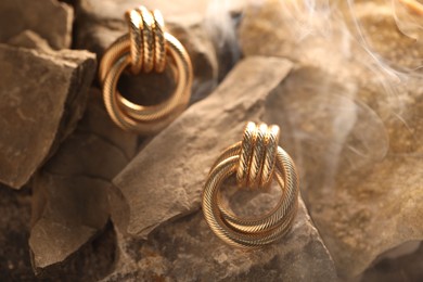 Photo of Stylish gold earrings in smoke on rocks, closeup